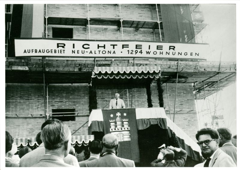 OÄAltona StÄum Große Bergstraße, Altona-Altstadt ZÄ2000 e HÄPolizeidirektion West, Mörkenstraße Sig Ot 39/5/46 quer Beschreibung: Blick auf Redner und Tribüne bei Richtfest. Außenaufnahme, Neu-Altona, Sanierung, Ritual, Einkaufszentrum. Im Vordergrund ein Mann im Anzug, meist zur Rednertribüne schauend, mit dem Rücken zum Fotografen. Die Tribüne ist geschmückt mit einer großen Fahne mit Hamburg-Wappen, schräg oben auch die Deutschland-Fahne, Symbol, Tuch, Girlande und einer geschwungenen Markise. Oben darüber befindet sich ein Schild mit der Aufschrift: "Richtfeier Aufbaugebiet Neu-Altona, 1294 Wohnungen." Im Hintergrund eine hohe Häuserreihe mit Baugerüst.